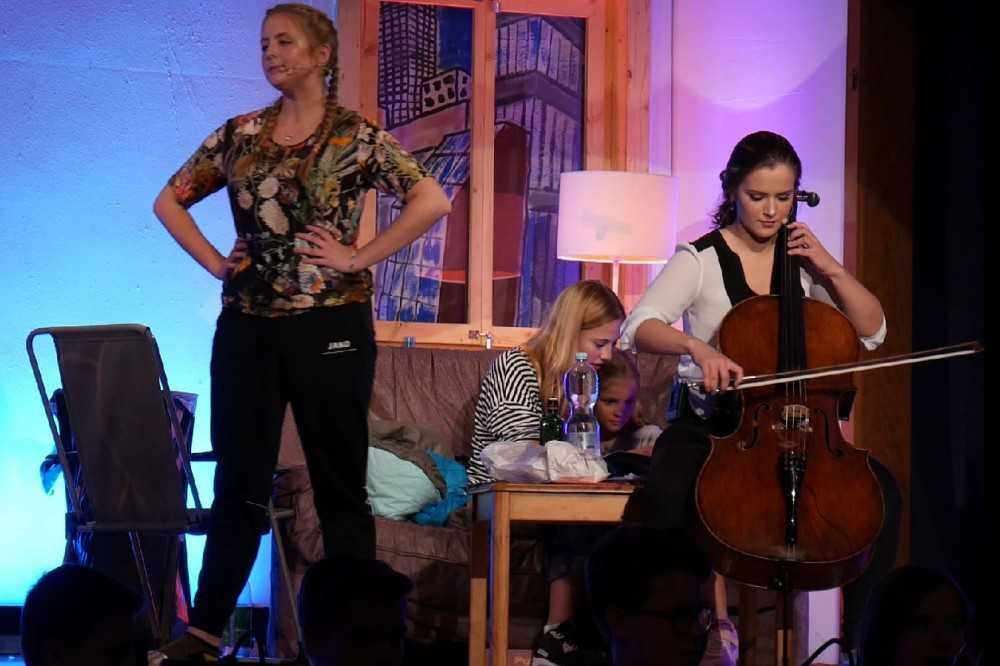 None - Links steht ein Mädchen mit den Händen in der Hüfte während rechts daneben ein Mädchen Cello spielt